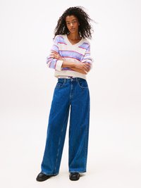 Kék, bő szabású farmernadrág, amelyet egy sokszínű pulóverrel viselnek, V nyakkal és rózsaszín és kék "Tommy Jeans" logóval.