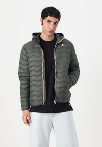 Green quilted puffer jacket with a hood, featuring zigzag stitching, grey zipper, and an embroidered logo at the chest. Paired with a black shirt.