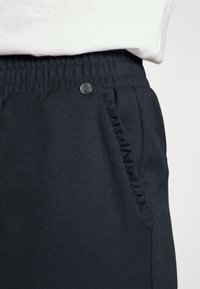 Svarta byxor med elastisk midja, med en framficka med volangkant och en metallknapp som accent. Slät tygtextur.