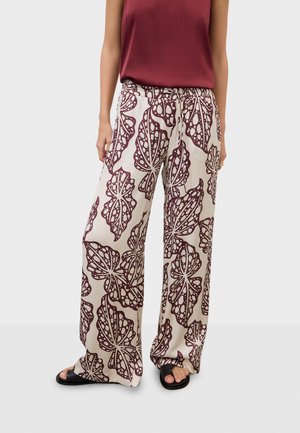 Kvinde iført beige bukser med vide ben og store mørkerøde bladmønstre, kombineret med en ærmeløs mørkerød top og sorte sandaler.