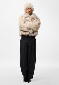 Krótka kurtka z futra ekologicznego w jasnobeżowych odcieniach z ciemniejszymi plamami, zestawiona z szerokimi czarnymi spodniami i pasującą czapką z tego samego futra ekologicznego.