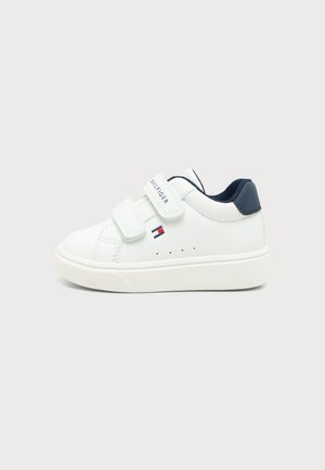 Baskets basses blanches avec deux bandes Velcro, languette talon bleu marine et petit logo drapeau rouge, blanc et bleu sur le côté.