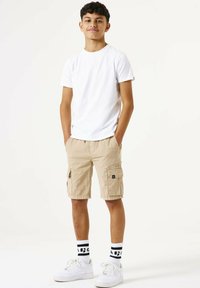 Witte T-shirt met korte mouwen, beige cargoshorts met zakken, zwart logo, gecombineerd met witte sneakers en zwart-wit gestreepte sokken.