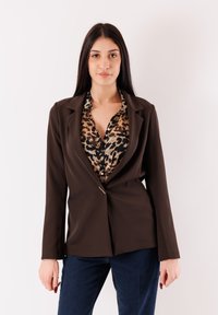 Blazer marrone sartoriale con collo sciallato, chiusura con un solo bottone e maniche lunghe, indossato sopra una camicia a stampa leopardata e jeans scuri.