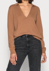 Kobieta w brązowym swetrze z dekoltem w serek i długim rękawem, wsunięta w czarne dżinsy z wysokim stanem, stoi z rękami swobodnie opadającymi wzdłuż ciała.