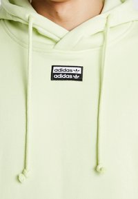 Světle zelená mikina s kapucí vyrobená z měkké tkaniny, která má nastavitelnou šňůrku a černou nášivku loga adidas na přední straně.