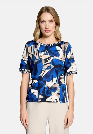 Vrouw met licht haar draagt een shirt met korte mouwen met een blauw, beige en zwart abstract bloemmotief en lichtgekleurde broek.