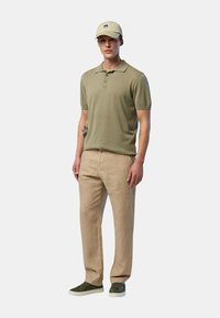 Polo shirt verde oliva, pantaloni beige e sneakers verdi. Il modello indossa un cappellino chiaro con un logo. Tessuto morbido, design casual.
