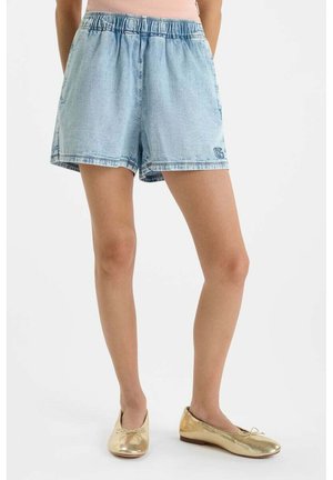Vrouw draagt lichtblauwe denim shorts met elastische tailleband en gouden ballerina's, staand tegen een witte achtergrond.