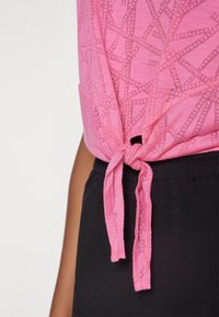 Top rosa con patrón y detalle anudado en el dobladillo, combinado con pantalones de cintura elástica negros. La tela es suave con una textura ligera.