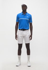 Homme debout portant un polo bleu, un short blanc, une casquette blanche, des chaussettes blanches et des baskets blanches sur un fond uni.