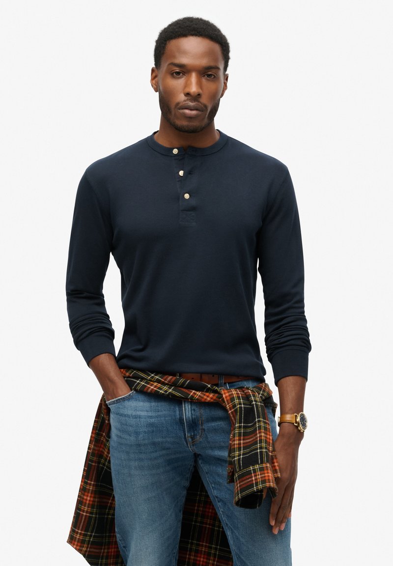 Marineblå langærmet henley skjorte med rund hals og tre knapper; parret med lyseblå jeans og en sort og orange ternet skjorte knyttet om taljen.