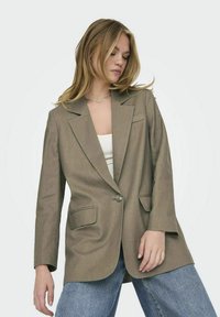 Blazer marrone su misura con rever a punte, chiusura con un bottone, due tasche frontali e una trama liscia, indossato sopra una maglietta bianca.