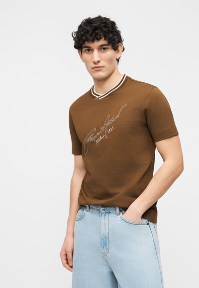 Νέος άνδρας με σγουρά σκούρα μαλλιά, φορώντας καφέ κοντομάνικο πουκάμισο με άσπρο κείμενο και ανοιχτόχρωμο μπλε τζιν, στέκεται μπροστά σε απλό φόντο.