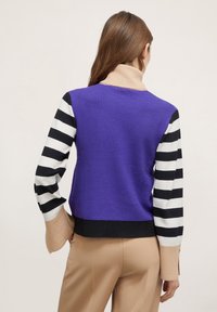 Fioletowy sweter wełniany z prążkowanymi czarno-białymi rękawami. Ma obcisły krój i ściągacz przy szyi. Jasnobrązowe akcenty przy mankietach.
