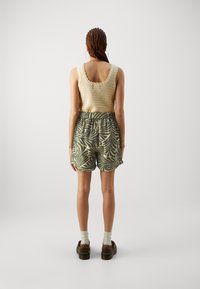 Pletený béžový top s vlnitými okraji, doplněný zelenými šortkami s listovým vzorem. Hnědé boty s bílými ponožkami dokončují outfit.