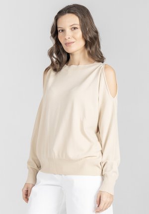 Kobieta o falujących brązowych włosach, ubrana w beżowy sweter z odsłoniętymi ramionami i białe spodnie, stojąca na jednolitym jasnoszarym tle.