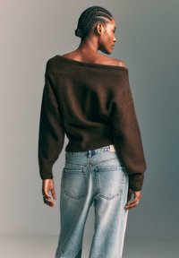 Bruine, oversized gebreide trui met off-shoulder ontwerp; gecombineerd met lichte, high-waisted jeans; zachte textuur en relaxed fit.