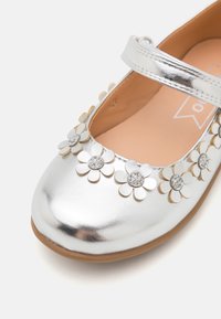 Stříbrné baleríny Mary Jane s hladkým metalickým povrchem, zdobené květinovými akcenty s kamínkovými středy a hnědou podešví.