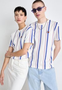 T-shirts blancs à rayures bleues et rouges, à manches courtes, avec un logo. Un modèle porte une jupe blanche, l'autre porte un jean bleu clair.