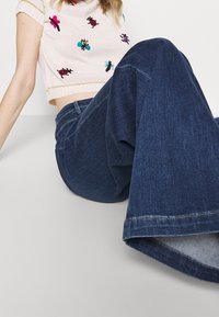 Vaqueros azules acampanados de cintura alta, hechos de denim, con una apertura de pierna ancha. Combinados con una blusa de manga corta bordada.
