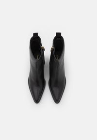 Botas negras de cuero hasta el tobillo con punta afilada, textura suave y acentos de cremallera dorada. Presentan una pestaña en la parte posterior para facilitar el uso.