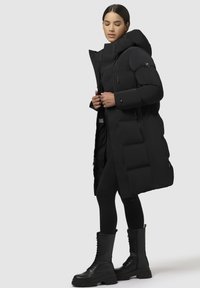 Melnā puffa jaka ar kapuci, ar kvadrātveida šuvēm, priekšējo rāvējslēdzēju un sānu kabatām. Kombinēta ar melnajām legingām un auklu zābakiem.