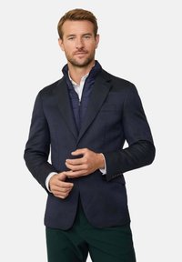 Blazer navy in tessuto testurizzato, colletto foderato a contrasto e un gilet imbottito con zip sotto. Camicia bianca e pantaloni verde scuro.