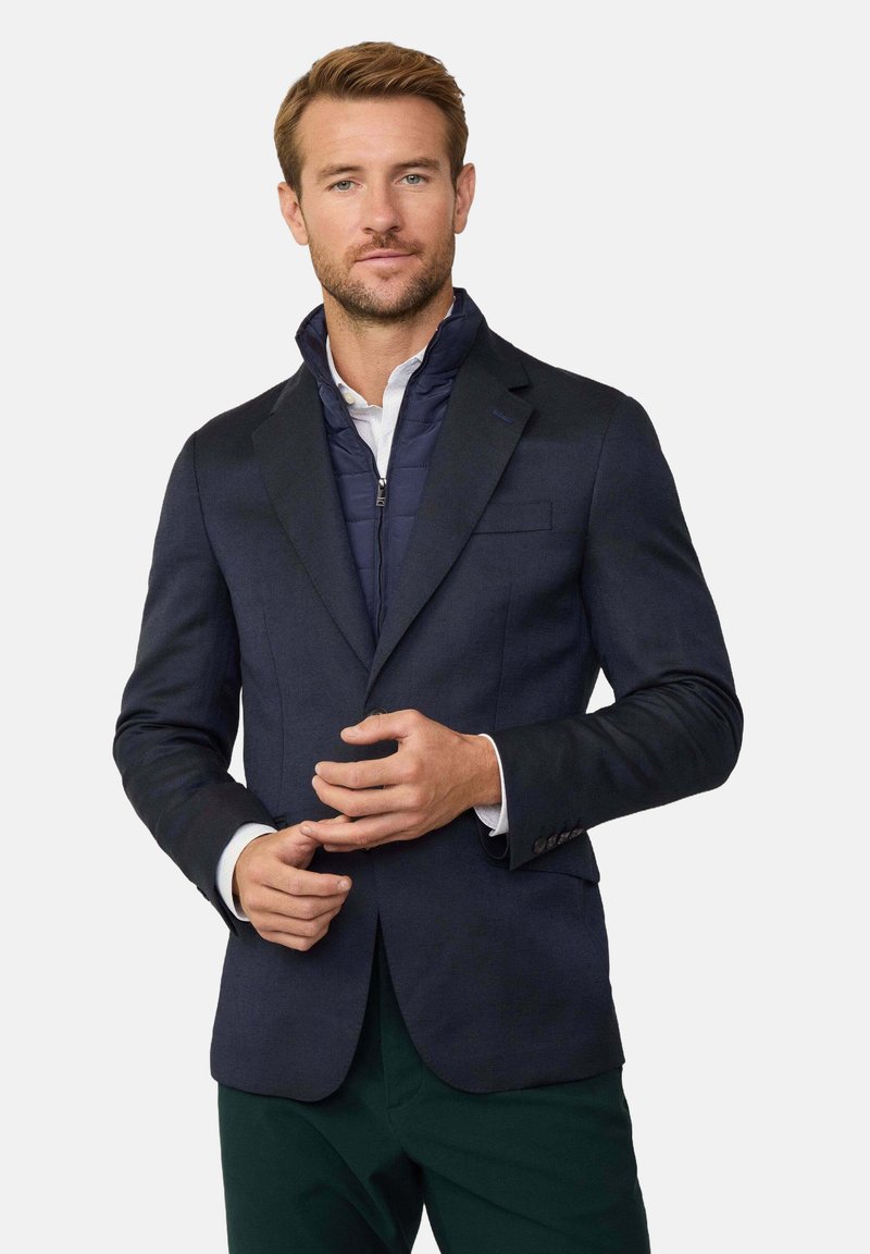 Blazer navy in tessuto testurizzato, colletto foderato a contrasto e un gilet imbottito con zip sotto. Camicia bianca e pantaloni verde scuro.
