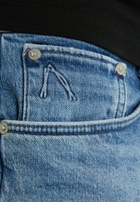 Šviesiai mėlyni džinsai su maža priekinė kišenė, sidabrinėmis detalėmis ir siuvinėta akcentų detale, primenančia "A" netoli kišenės.