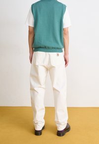 Persoon in een witte broek met achterzakken, een teal mouwloze sweater over een witte T-shirt met korte mouwen, en zwarte schoenen, staand op een gele vloer.