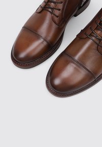 Zapatos de vestir de cuero marrón con un acabado pulido, punta redonda y diseño con cordones. Presentan costuras en contraste y un tacón ligeramente elevado.