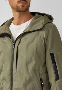 Olive zielona kurtka z kapturem o pikowanym wykończeniu, wyposażona w czarny zamek błyskawiczny, siateczkowe akcenty oraz boczne kieszenie. Gładka faktura z minimalnym wzorem.