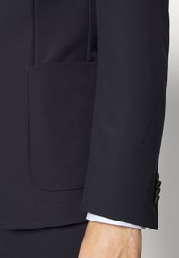 Marinblå kavaj med sidoficka, mjuk textur, skräddarsydd passform och knäppning vid ärmsluten. Tygerna verkar lätta och polerade.