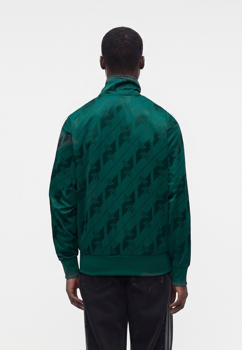 Hombre de espaldas que lleva una chaqueta verde oscuro con estampado y vaqueros negros sobre un fondo blanco liso.