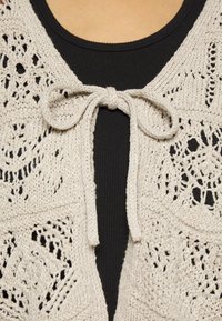 Knit cardigan vaaleanbeigessä sävyssä, jossa on pitsikuvioita ja etuosassa narukiinnitys. Pukeutuneena mustan paidan päälle, paljastaen teksturoitua kangasta.