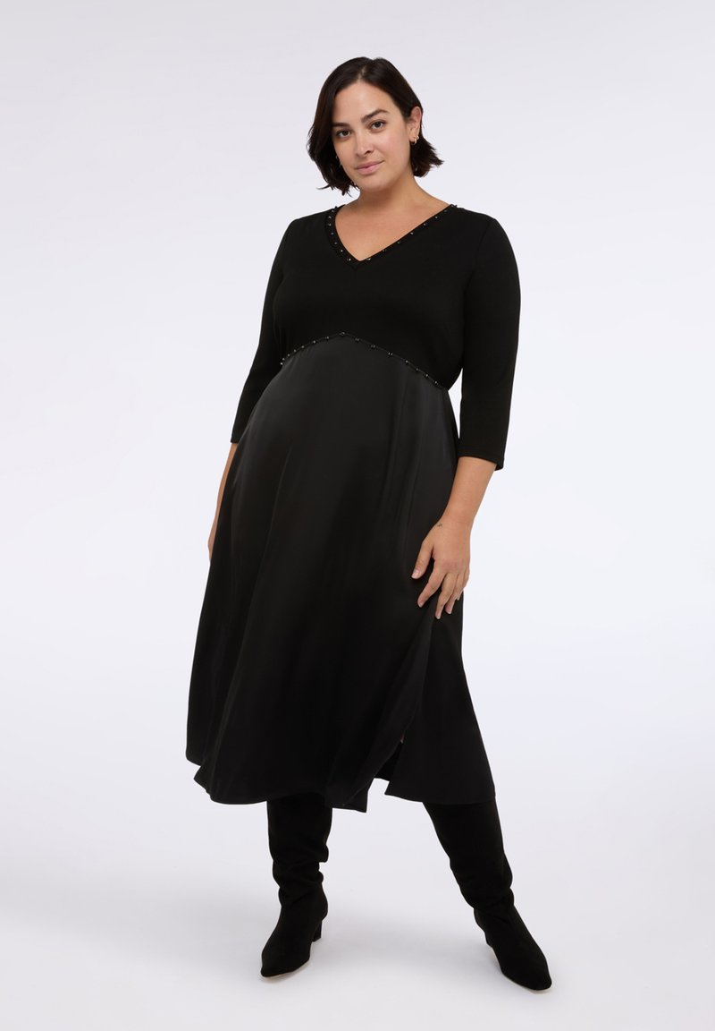 Černé šaty s výstřihem do V a tříčtvrtečními rukávy, s hladkou saténovou sukní a dekoračními nýty podél pasu.