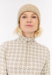 Beige a krémový top s vzorem houndstooth, vysokým límcem a krátkým zipem. Světle hnědá pletená čepice s ribbed vzorem sedí na vlnitých vlasech.