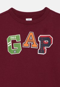 T-shirt en coton bordeaux avec un design graphique comprenant "1969" en vert, "GAP" en orange, "Athletic" et "NEW YORK" en lettres gras multicolores.