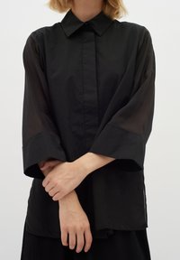 Osoba ubrana w czarn�ą bluzkę z długim rękawem i przezroczystymi rękawami, stojąca z założonymi rękami na przodzie, na jednolitym białym tle.