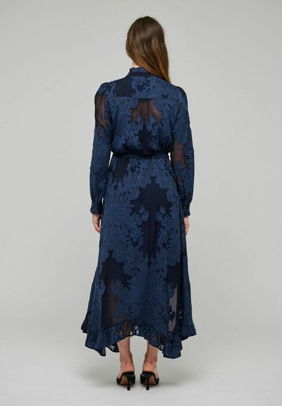 Femme portant une robe en dentelle florale bleu marine à manches longues, avec un col montant et un ourlet à volants, debout dos à la caméra.