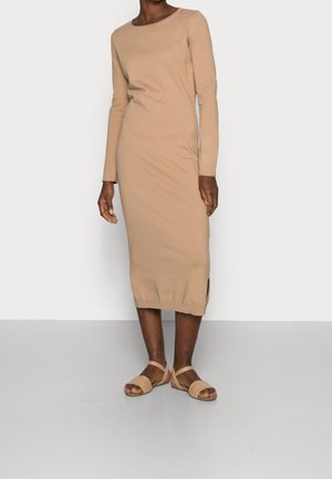 Abito a maglia beige a maniche lunghe con spacchi laterali, scollatura rotonda e silhouette aderente. Abbinato a sandali beige con cinturini.