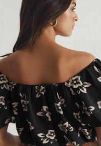 Zwarte off-the-shoulder top met een bloemenpatroon in roze en wit. Getextureerde stof met een gathered halslijn en korte mouwen.