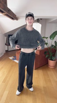 Pullover a righe bianche e nere, pantaloni della tuta neri e sneakers bianche. Ambiente interno con una pianta e un divano arancione.