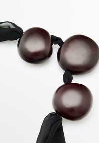 Perle di forma organica di un borgogna scuro collegate da un nastro di stoffa nera e testurizzata. Superficie liscia con una leggera lucentezza e bordi arrotondati.