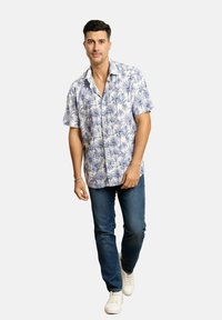 Camisa de manga corta de color claro con un patrón de palmeras azules, combinada con jeans oscuros y zapatillas blancas. Ajuste casual y diseño abotonado.