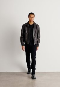 Chaqueta bomber de cuero negro con cremallera en el frente, llevada sobre una camiseta negra y jeans ajustados negros. El modelo lleva botas negras hasta el tobillo.