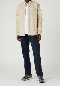 Camicia beige con bottoni, taglio regolare, con taschino sul petto, indossata sopra una t-shirt bianca e abbinata a pantaloni color navy e sneaker grigie.