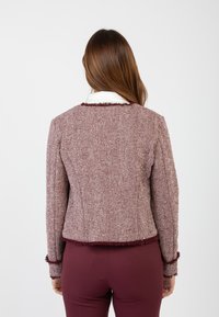 Giacca bordeaux con trama a spina di pesce e dettagli a frange sui bordi, con un taglio su misura, indossata sopra pantaloni rossi attillati.
