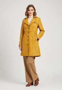 Yellow leather trench coat with a tailored fit, pointed collar, button closure, and two side pockets, paired with beige wide-leg trousers.